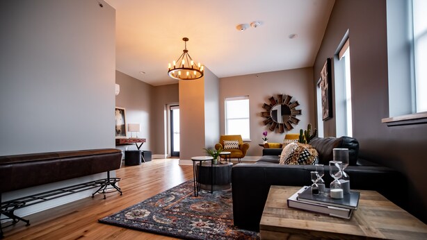 A spacious living room with a wooden floor, a black sofa, a coffee table with books and hourglasses, a yellow armchair, and a large wall clock. - Duets Bed & Breakfast (Tulsa)