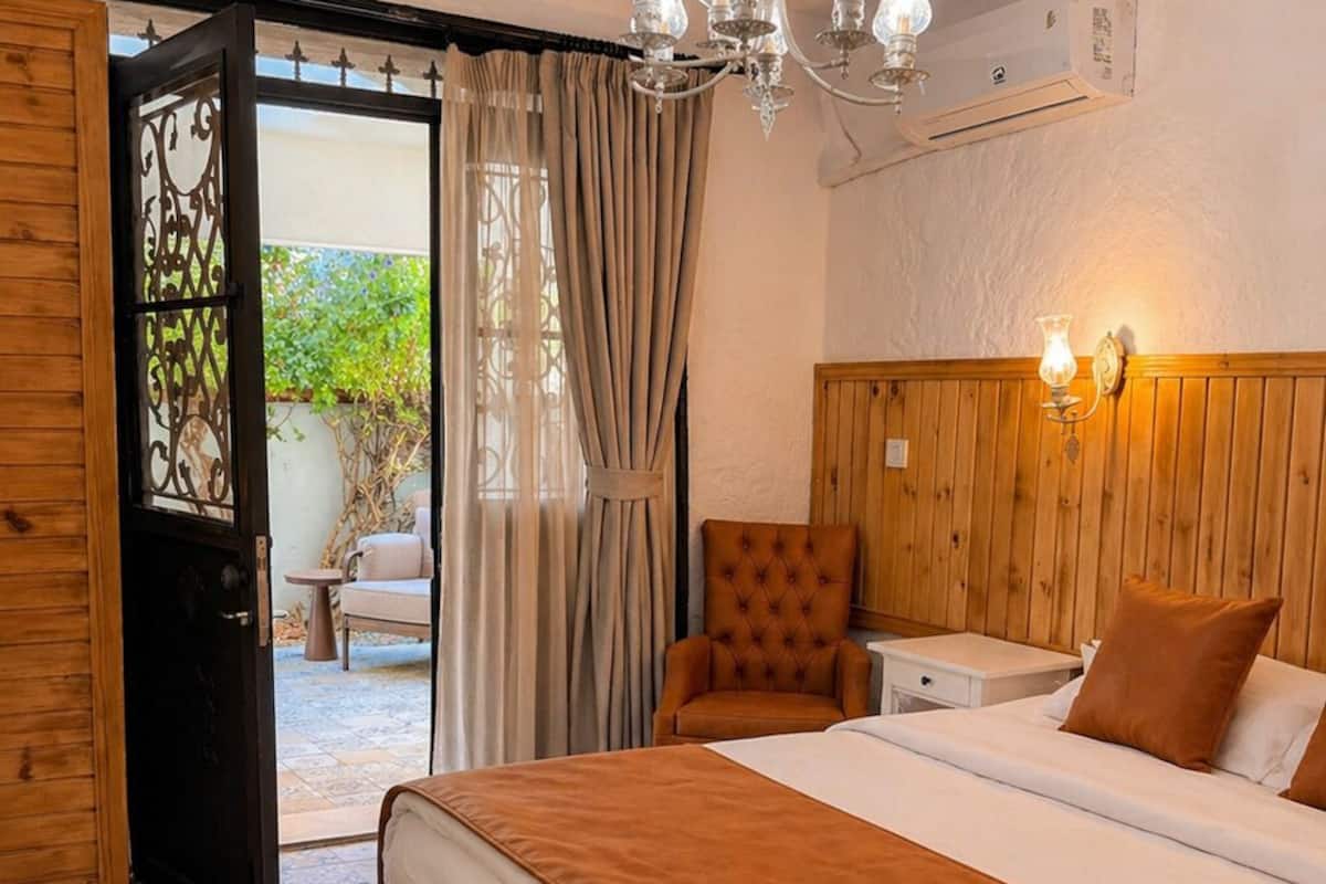 A hotel room with a bed, a chair, a nightstand, a chandelier, and an open door leading to an outdoor area.