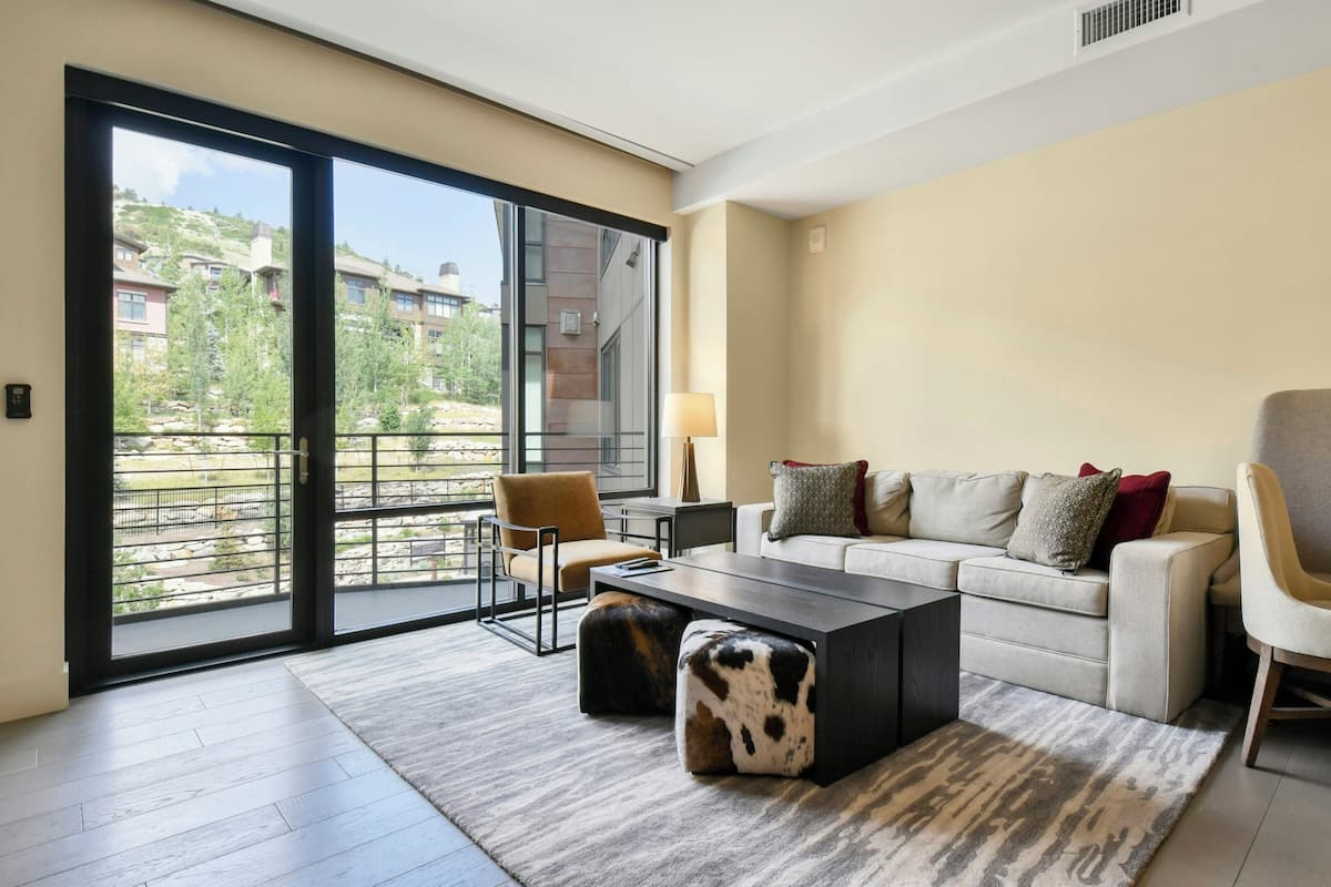 Una sala moderna con sofá, sillón y reposapiés, una mesa de centro, y vistas a un paisaje montañoso a través de amplios ventanales correderas.