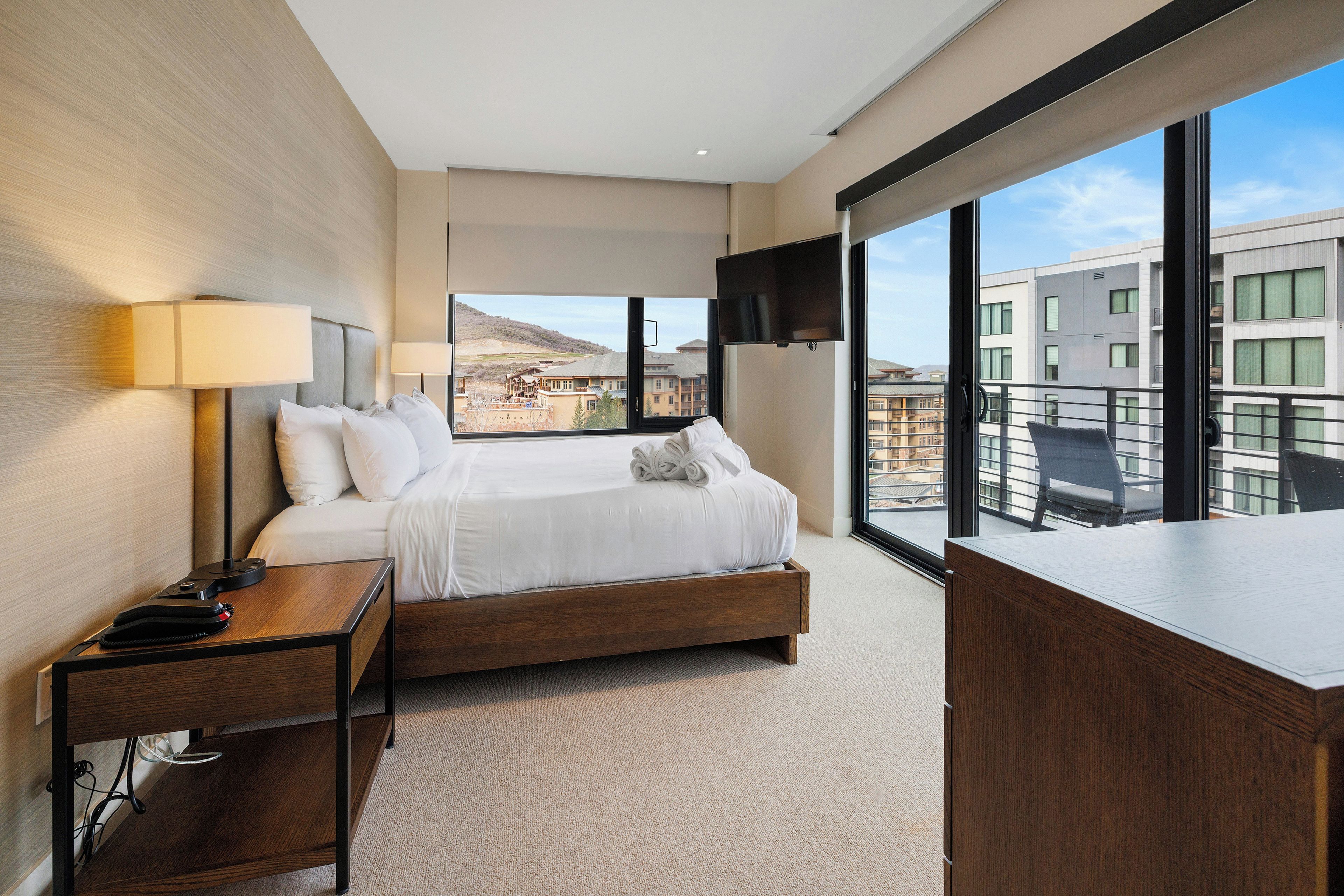 Habitación de hotel con una cama grande, un escritorio, un televisor y un balcón con vista a la ciudad.
