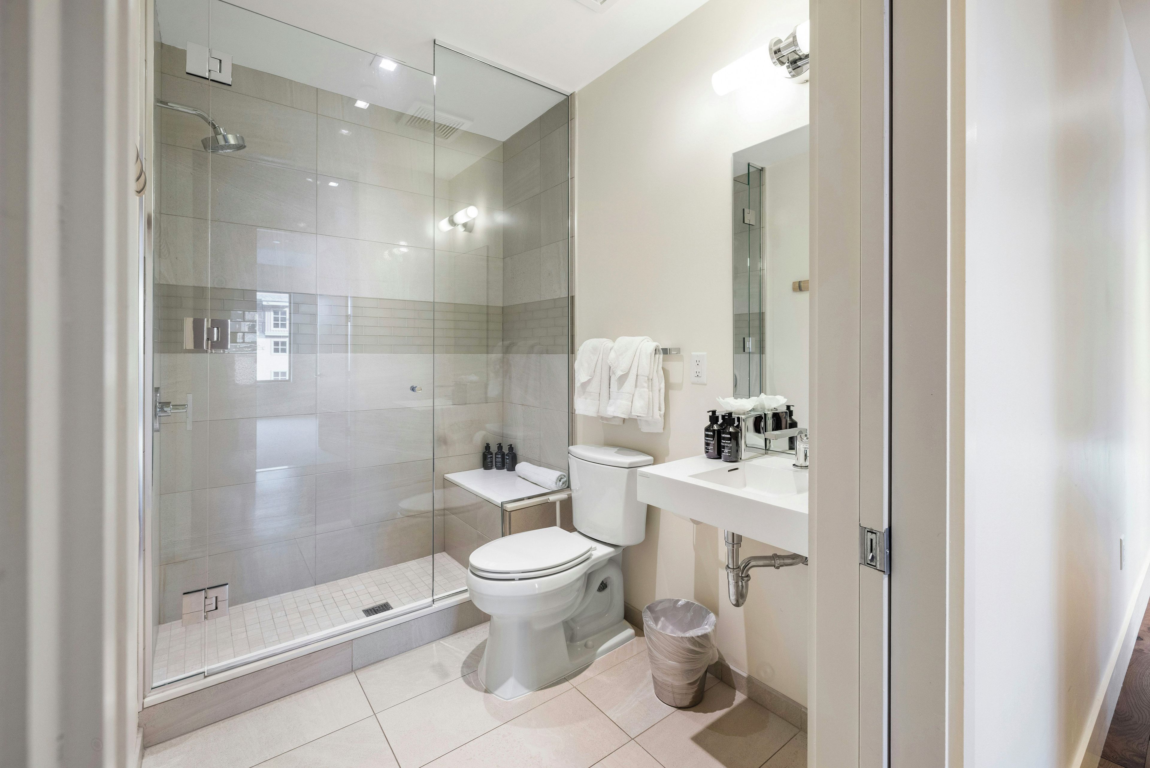 Un baño moderno con ducha envuelta en vidrio, inodoro, lavabo con artículos de aseo y un cubo de basura.