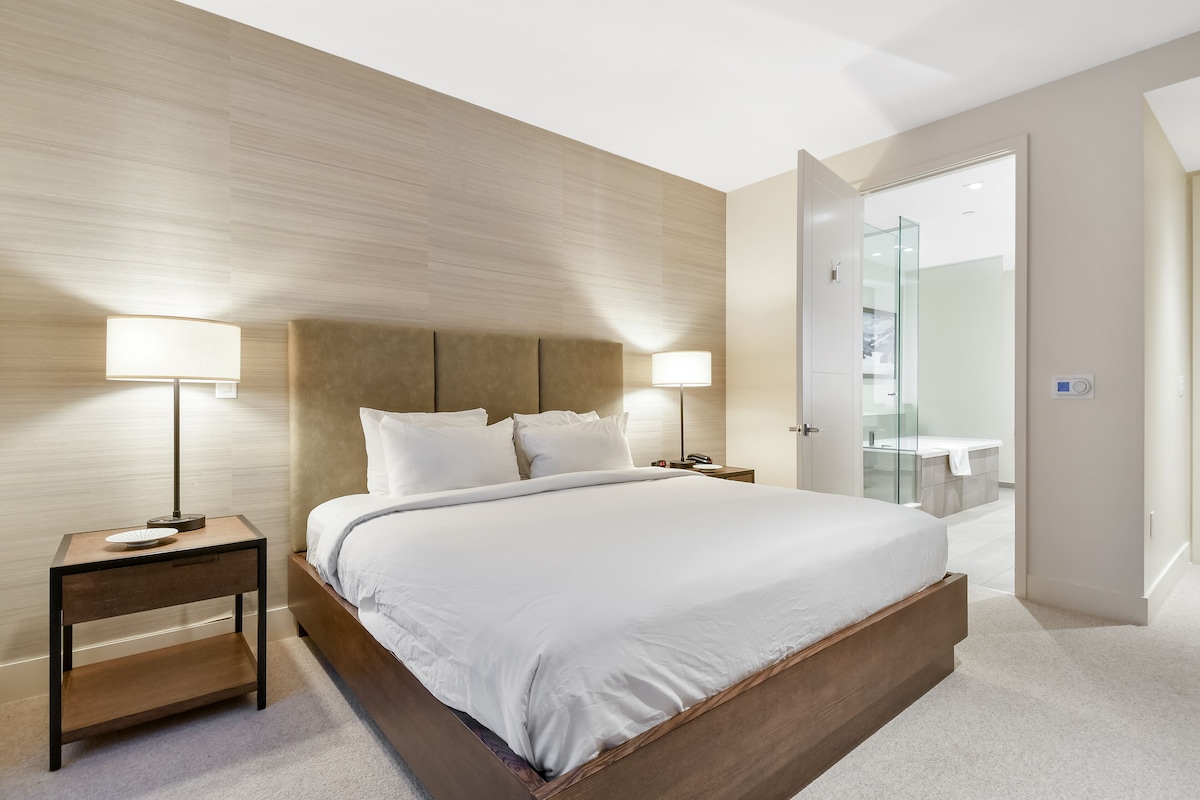 Une chambre moderne avec un grand lit, deux tables de chevet avec des lampes, une tête de lit en bois et une cabine de douche en verre.