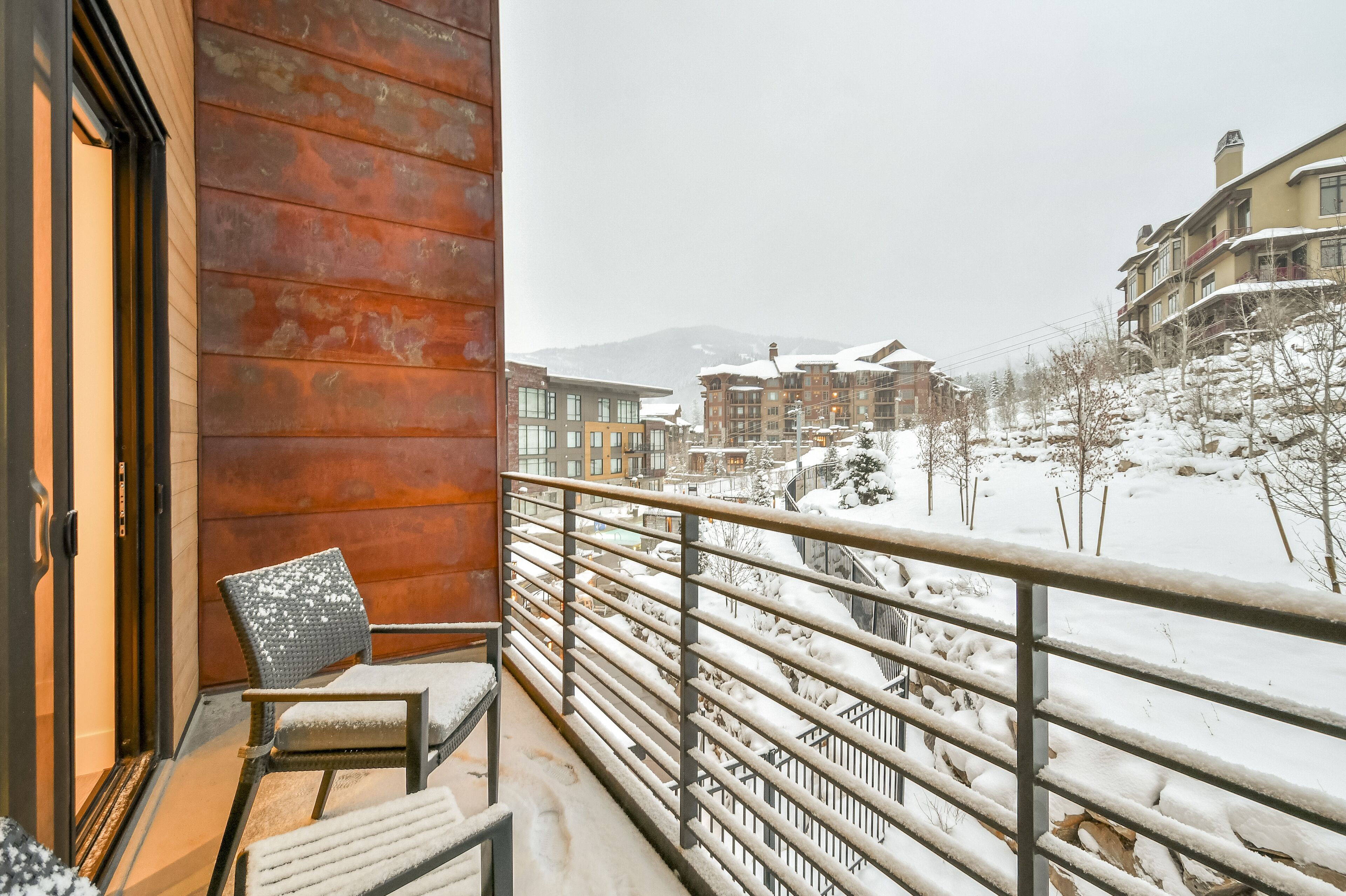 Un balcon avec une chaise, une rambarde en métal et une vue sur des bâtiments et des arbres recouverts de neige.