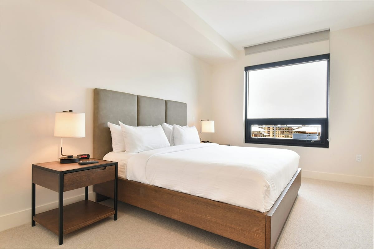 Una cama bien tendida con estructura de madera y ropa de cama blanca, una mesita de noche con lámpara, y una ventana con vistas a edificios.