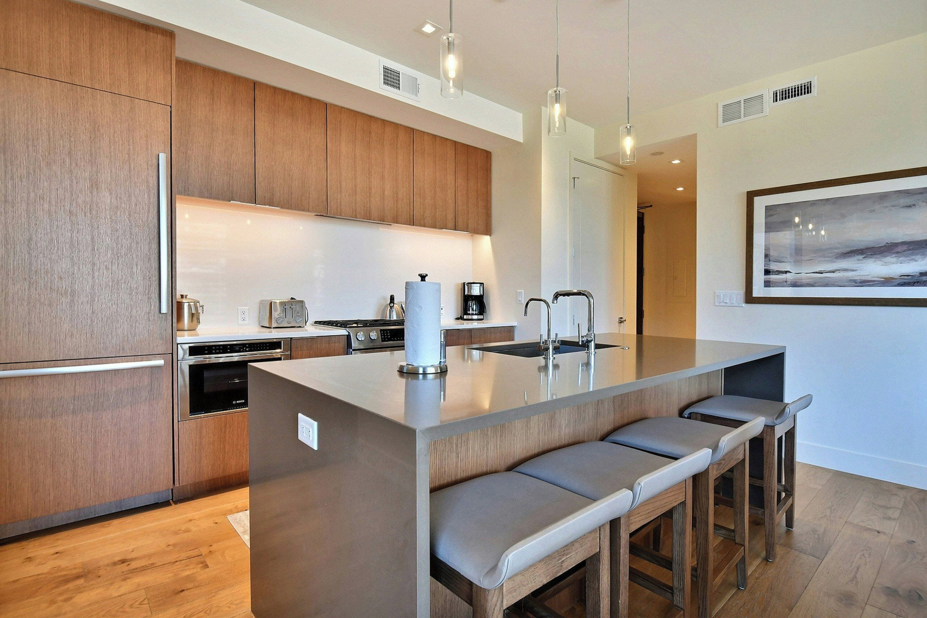 A modern kitchen with wooden cabinets, a central island with bar stools, stainless steel appliances, and pendant lighting.