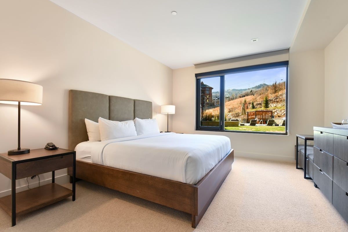 Un hotel moderno con una cama grande, una mesita de noche, un escritorio con lámpara y vistas a un paisaje montañoso a través de un ventanal amplio.