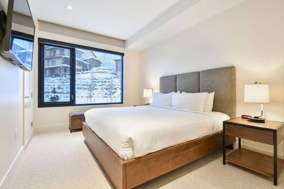 Un dormitorio moderno con una cama grande, mesitas de noche, un ventanal con vistas a un paisaje nevado y un televisor montado.