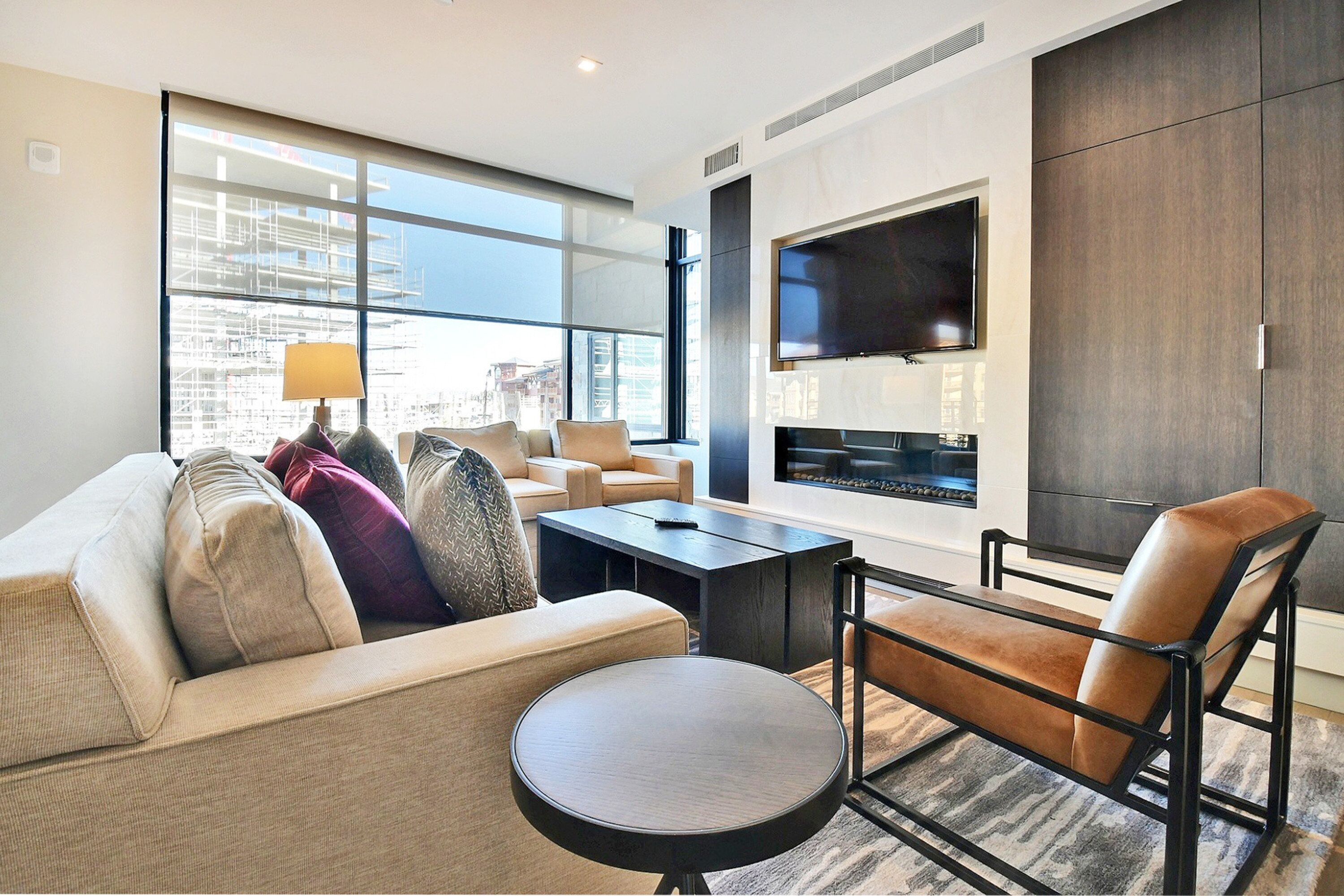Una sala moderna con un sofá, un sillón y una mesa de centro. Un televisor de pantalla plana está montado en la pared, y hay una ventana con vistas a edificios.