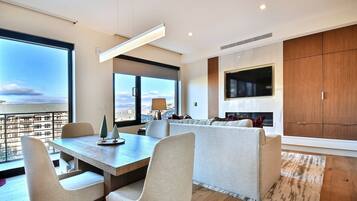A modern living room with a dining area, large windows, a fireplace, and a TV mounted on the wall.