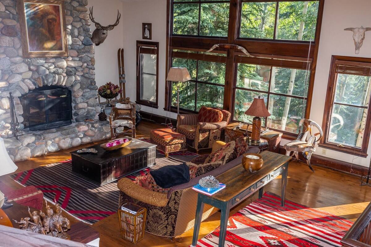 A rustic interior with a stone fireplace, wooden furniture, and large windows offering a view of greenery.