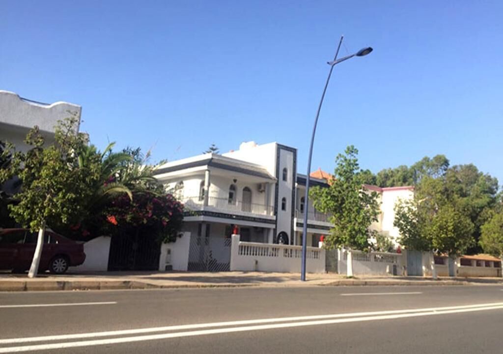 Un edificio de dos pisos con fachada blanca y techo plano, situado en una calle bajo un cielo despejado.