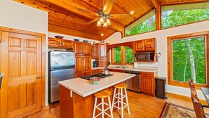 A spacious kitchen with wooden cabinets, a large island with stools, a ceiling fan, and a view of trees through a large window. - Mountain Lure: VIEWS OF BLUE RIDGE MTNS & LAKE LURE - NEAR ASHEVILLE & BLACK MTN (Old Fort)
