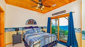 A bedroom with a bed, a nightstand, a ceiling fan, a window with curtains, and a view of the outdoors. - Mountain Lure: VIEWS OF BLUE RIDGE MTNS & LAKE LURE - NEAR ASHEVILLE & BLACK MTN (Old Fort)