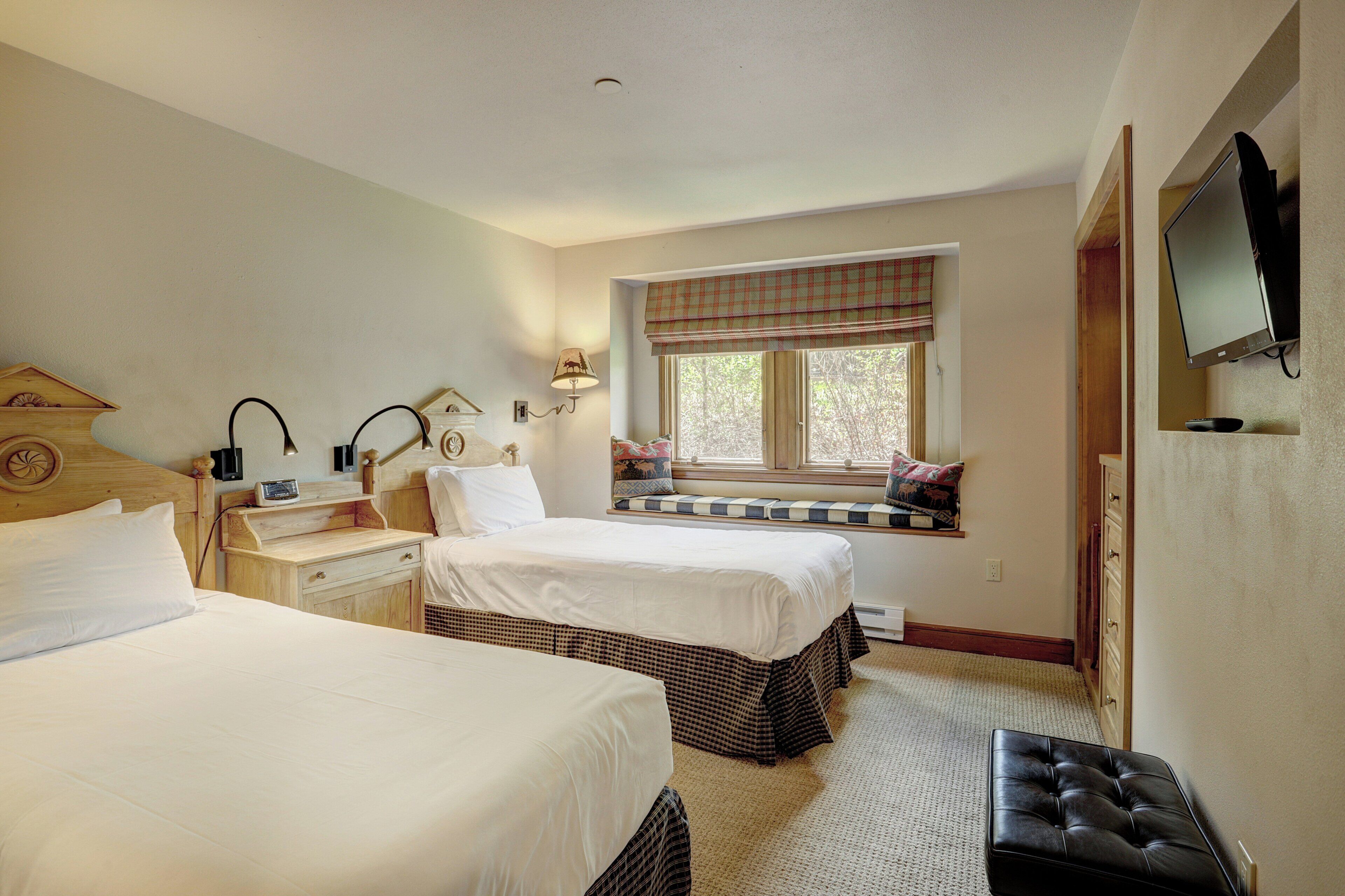 A hotel room with two beds, a window with a checkered blind, a television mounted on the wall, and a small table with a lamp.