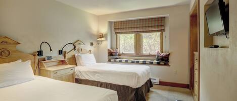 A hotel room with two beds, a window with a checkered blind, a television mounted on the wall, and a small table with a lamp.