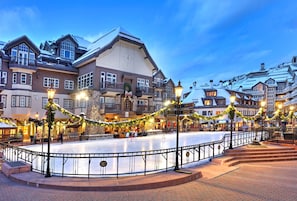 A well-lit plaza with a central ice rink, surrounded by multi-story buildings with balconies and outdoor seating areas.
