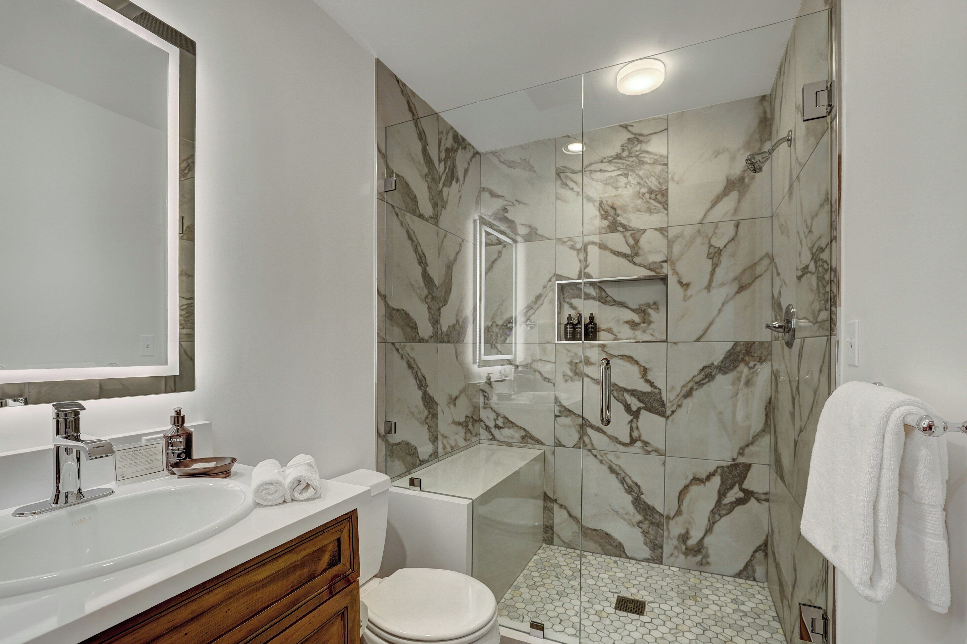 Een moderne badkamer met een grote spiegel, een wit wastafelmeubel, een douche met marmeren tegels en een toilet.