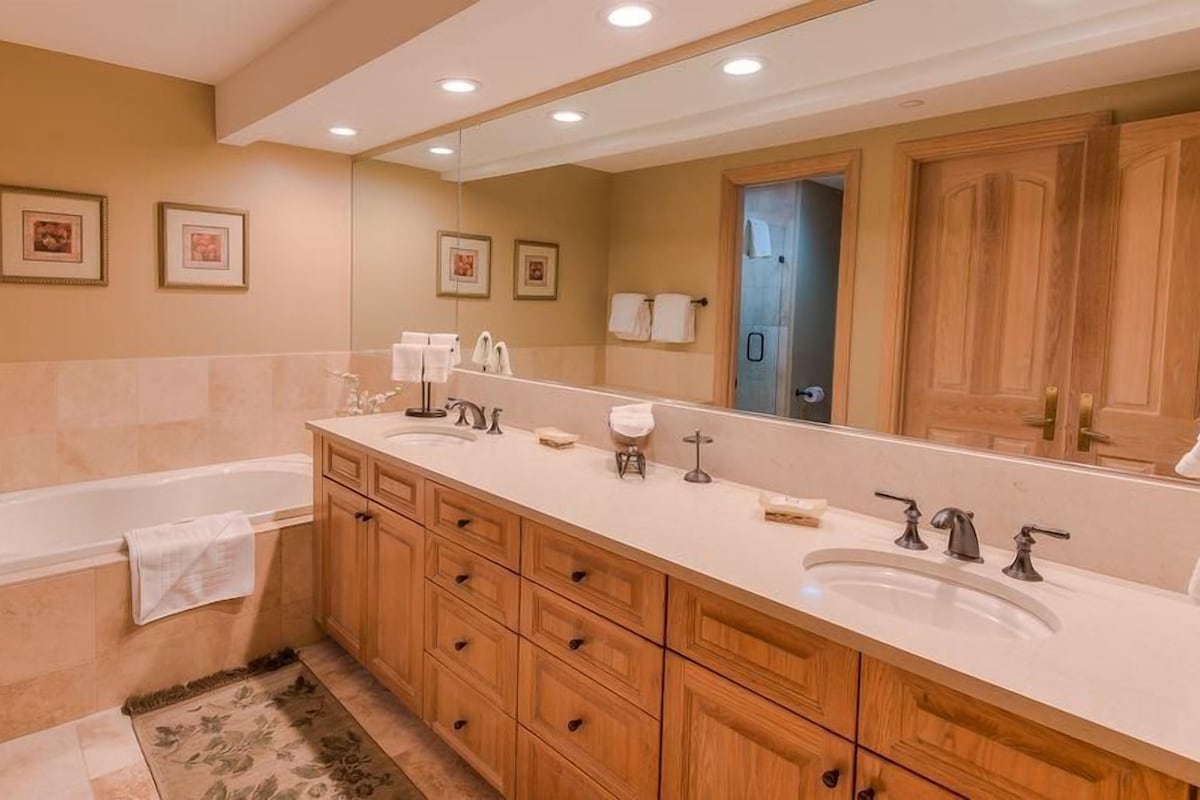 A bathroom with a bathtub, two sinks, wooden cabinets, a large mirror, and framed pictures on the wall.