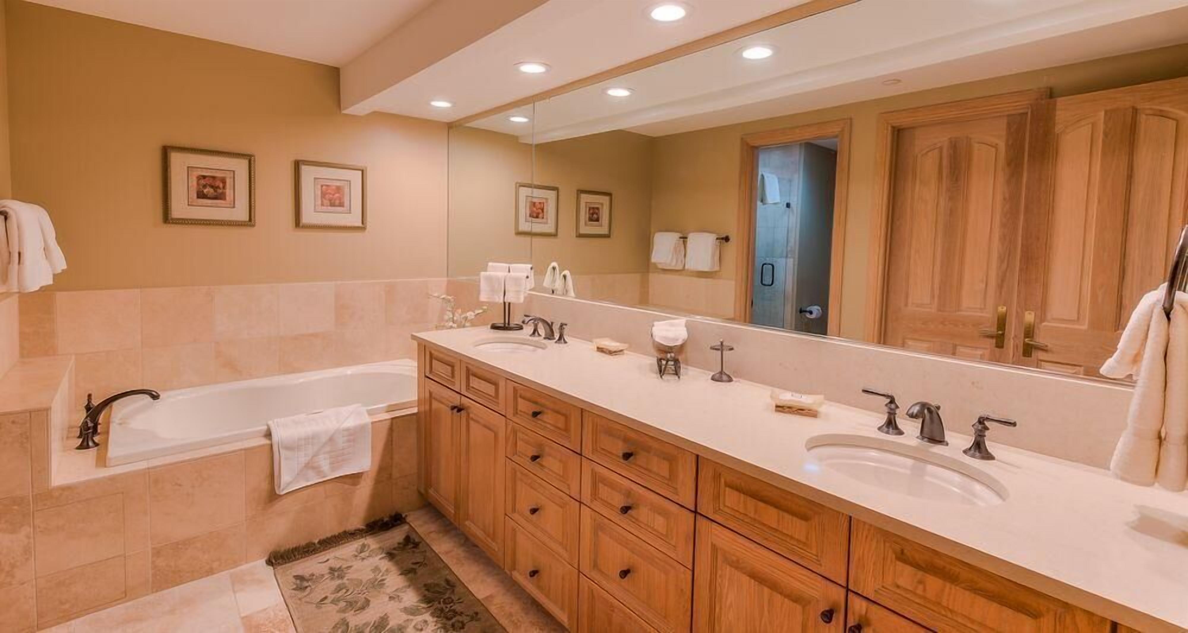 A bathroom with a bathtub, two sinks, wooden cabinets, a large mirror, and framed pictures on the wall.