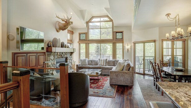 A spacious living room with a large window, a fireplace, and a dining area with a high ceiling and chandelier. - Beaver Creek Village by Vail Resorts (Avon)