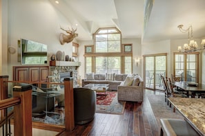 A spacious living room with a large window, a fireplace, and a dining area with a high ceiling and chandelier. - Beaver Creek Village by Vail Resorts (Avon)