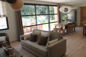 A modern living room with a sofa, dining table, and chairs. Large windows offer a view of the outdoors. - Gite Le Grand Angle (Mornas)