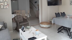 Ein gemütliches Wohnzimmer mit einer Couch, einem Esstisch mit Stühlen, einem an der Wand montierten Fernseher und einer Treppe, die in ein weiteres Zimmer führt.