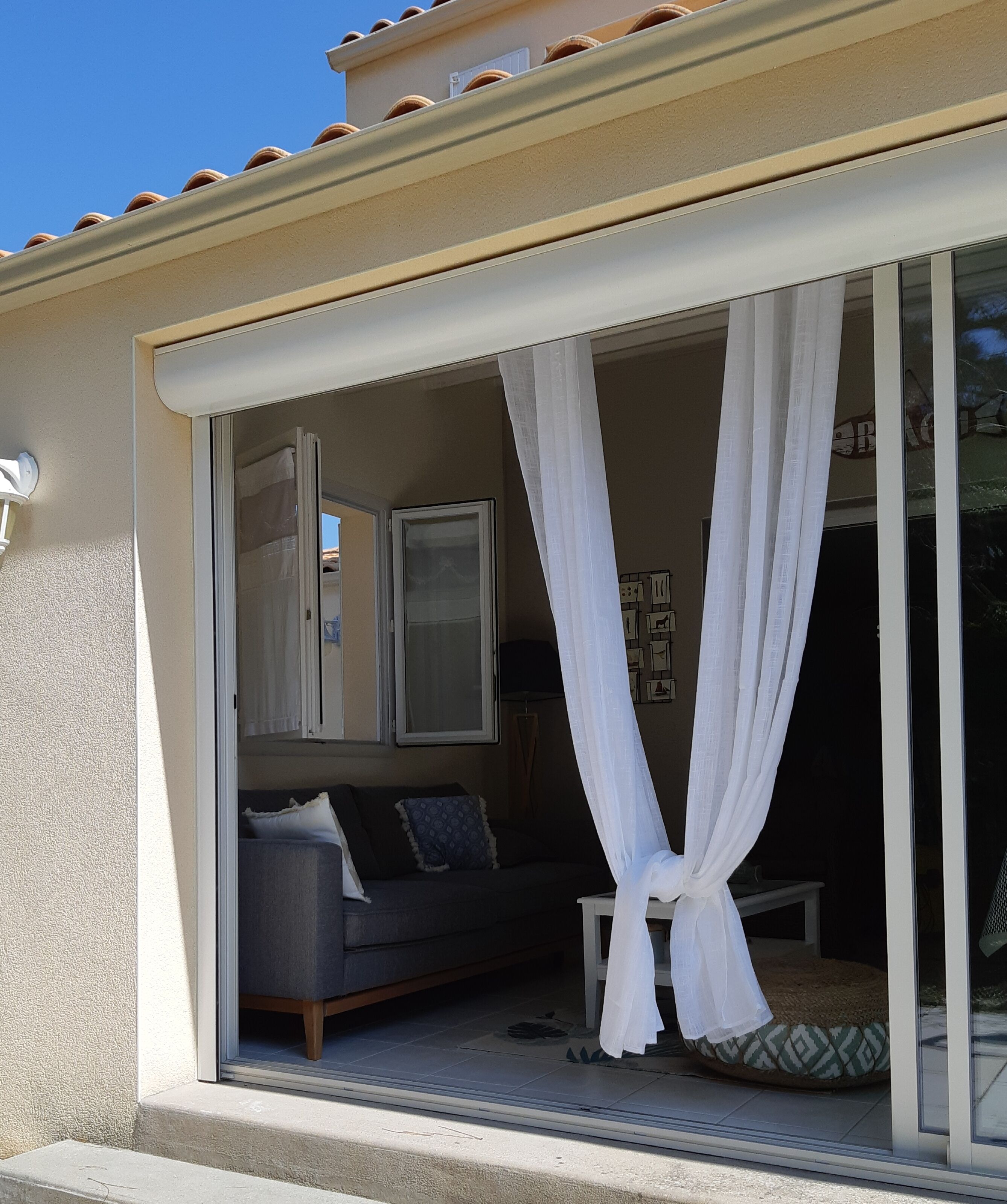 Quarto com porta de vidro deslizante, sofá cinza e cortina branca.
