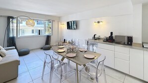 A modern kitchen with a dining area, clear chairs, a sofa, and a TV mounted on the wall.