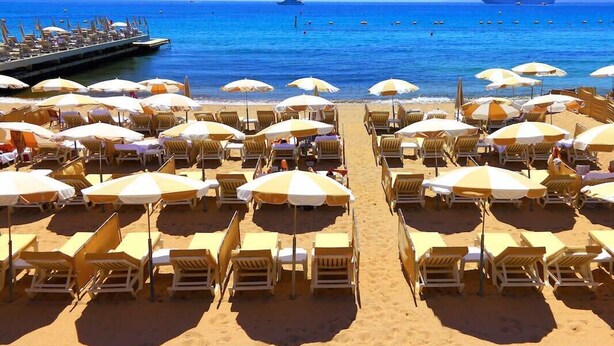 A beach with rows of sun loungers and umbrellas, a pier extending into the water, and a clear blue sky.