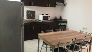 Een kleine keuken met donkere kastjes, een magnetron, een waterkoker en een eettafel met stoelen.