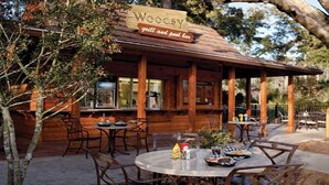 Een rustieke houten buitenzitplaats met tafels en stoelen, een bord met de tekst 'Woody grill and pool bar' en uitzicht op de omliggende bomen.