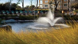 Een fontein in een aangelegde tuin met een zwembad, omringd door gras en bomen.
