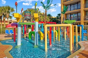 A water park with colorful play structures, a pool, and a building with balconies in the background.