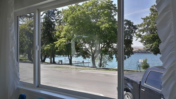 A view through a window of a parking area with a body of water and trees in the background. - Stunning Lake Views from Every Room! (Polson)