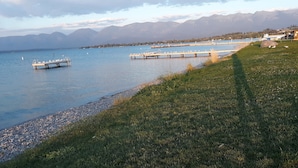 A lakeside view with a dock extending into the water, surrounded by mountains and a grassy area. - Stunning Lake Views from Every Room! (Polson)