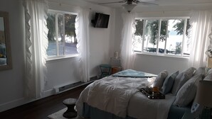 A bedroom with a bed, a ceiling fan, two windows with curtains, a small table, and a chair. - Stunning Lake Views from Every Room! (Polson)