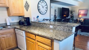 Eine Küche mit Granit Arbeitsplatten, Holzschränken, einem Geschirrspüler, einer Uhr und einem Essbereich mit Fernseher und einer Couch.