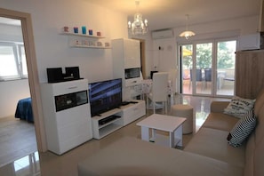 A modern living room with a flat-screen TV, white furniture, a chandelier, and a balcony with outdoor furniture.