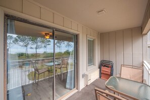 Un balcón con mesa y sillas, vistas a árboles, y una ventana con persianas.