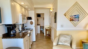 A modern kitchen with white cabinetry, a coffee maker, and a trash bin. There is a beige armchair and a small table with a lamp.