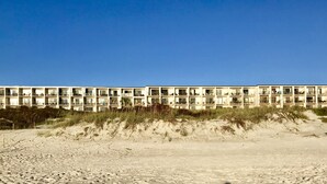 A long, multi-story hotel with balconies, situated on a sandy beach with dunes in the background.