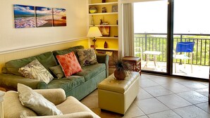 A living room with a green sofa, a beige ottoman, and a blue chair. There is a balcony with a view of greenery.