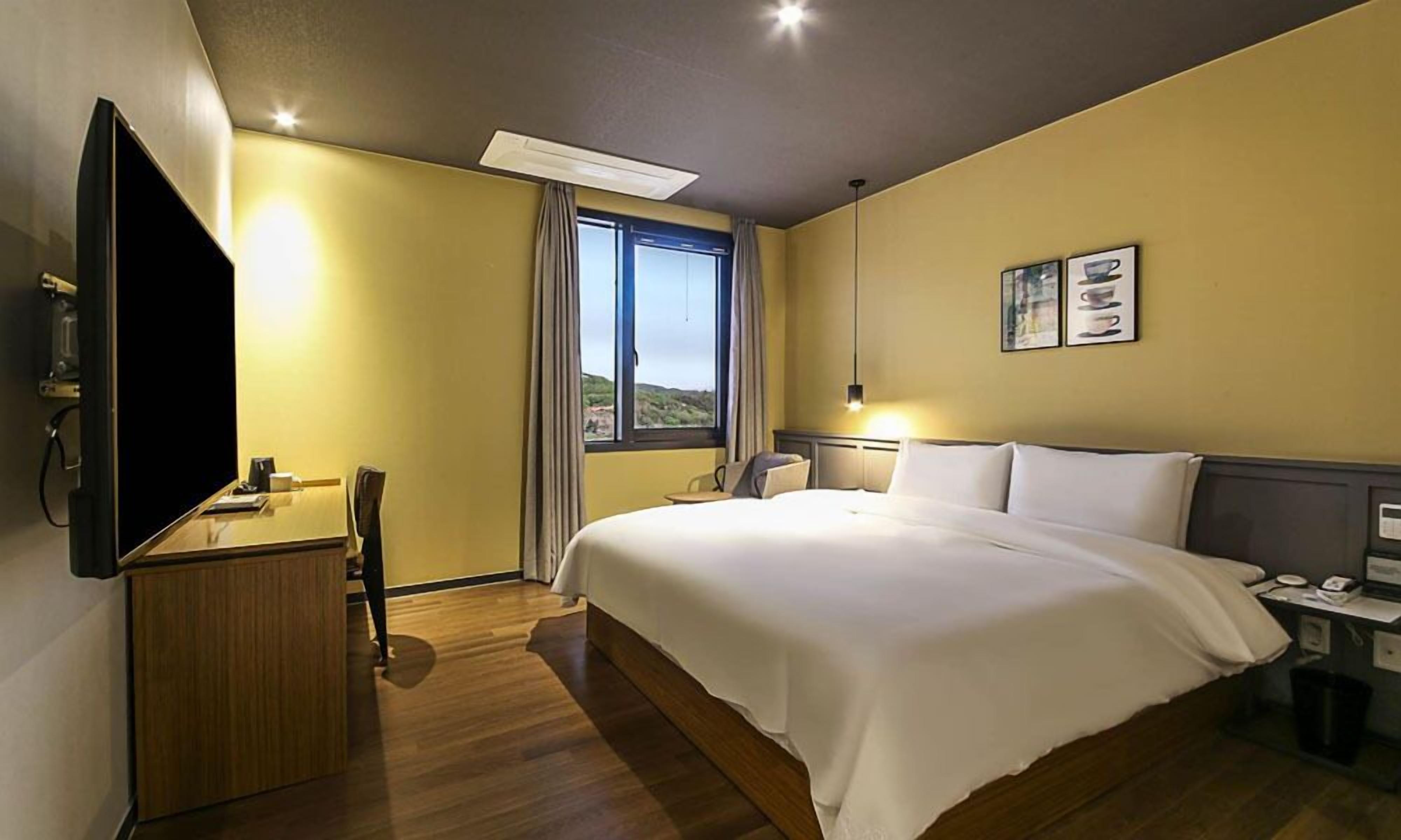 A hotel room with a bed, bedside table, lamp, TV, and a window with curtains.