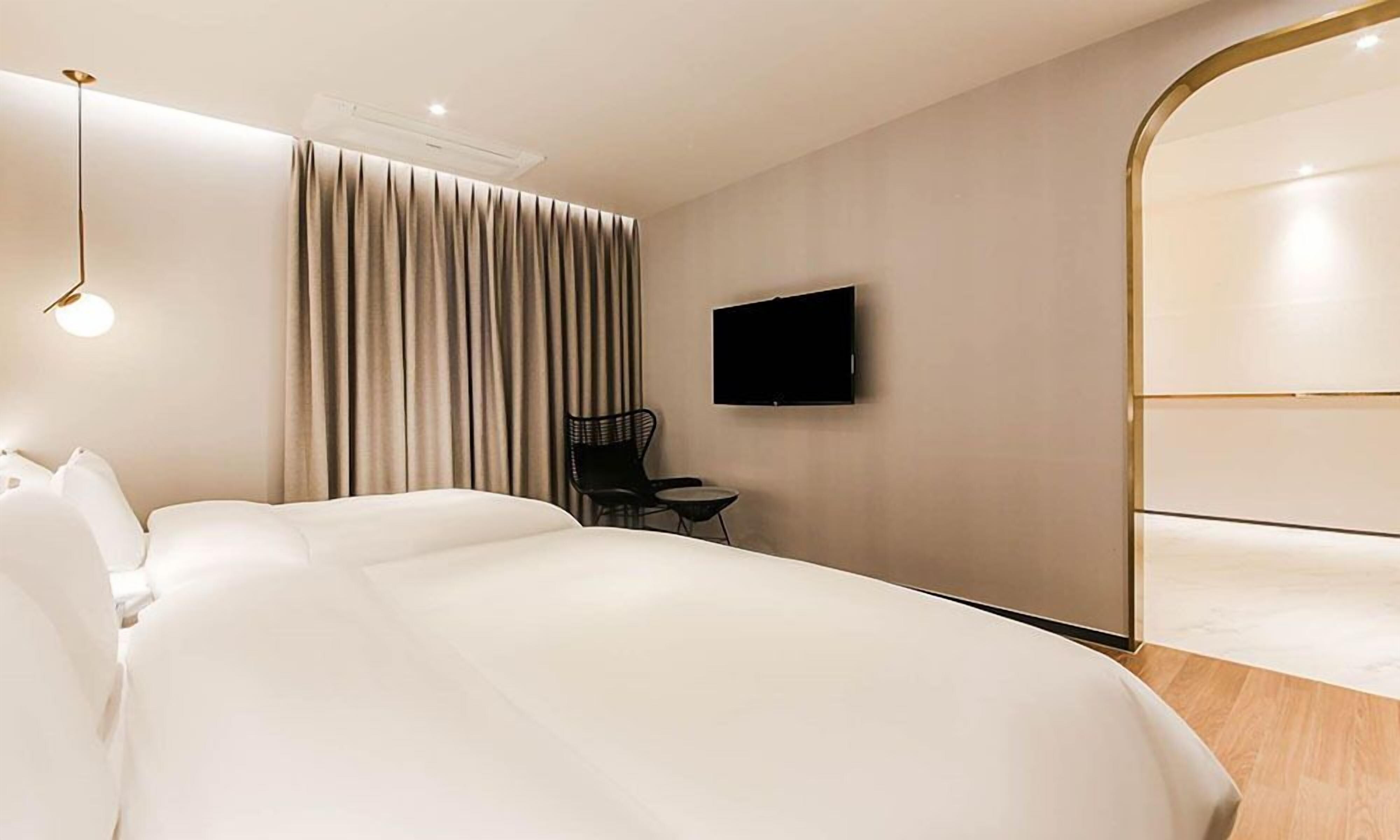 A modern hotel room with a large bed, a flat-screen TV mounted on the wall, a small table with a chair, and a large mirror.