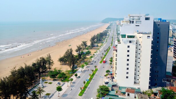 A coastal cityscape with a beach, a road lined with trees, and a tall building with multiple floors. - Phoenix Hotel 2 (Sam Son)