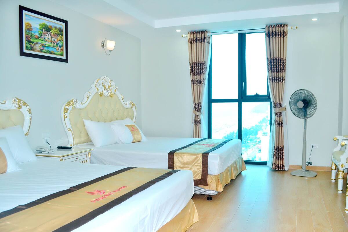 Une chambre d’hôtel avec deux lits, une grande fenêtre avec des rideaux, un ventilateur fixé au mur et un tableau encadré accroché au mur.