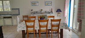 Ein Essbereich mit einem für sechs Personen gedeckten Tisch, umgeben von Holzstühlen mit floralem Bezug. Im Hintergrund ist eine Küche mit weißen Schränken und einem Fenster erkennbar.