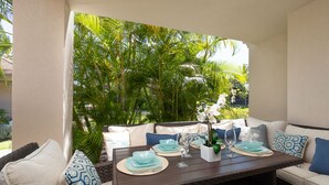 An outdoor dining area with a wicker table set for two, surrounded by cushioned seating and a backdrop of lush tropical foliage. - 🏖️2 Cal King Townhome | Pickleball + Pools | Near A-Bay (Waikoloa)