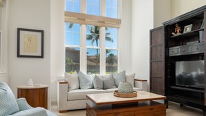 A living room with a sofa, coffee table, TV, and a window with a view of palm trees. - 🏖️2 Cal King Townhome | Pickleball + Pools | Near A-Bay (Waikoloa)